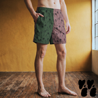 Model wearing mid-length swim trunks featuring a split olive green and dusty pink colorway with black 1950s tiki girls, palm trees, and tiki totem illustrations.