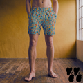 Model wearing mid-length swim trunks with a mid-century geometric mosaic tile pattern in teal blue, terracotta orange, mustard yellow, and muted pink.