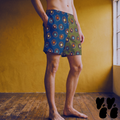 Model wearing mid-length swim trunks with hand-drawn retro vinyl record illustrations on a split blue-green and olive green background.