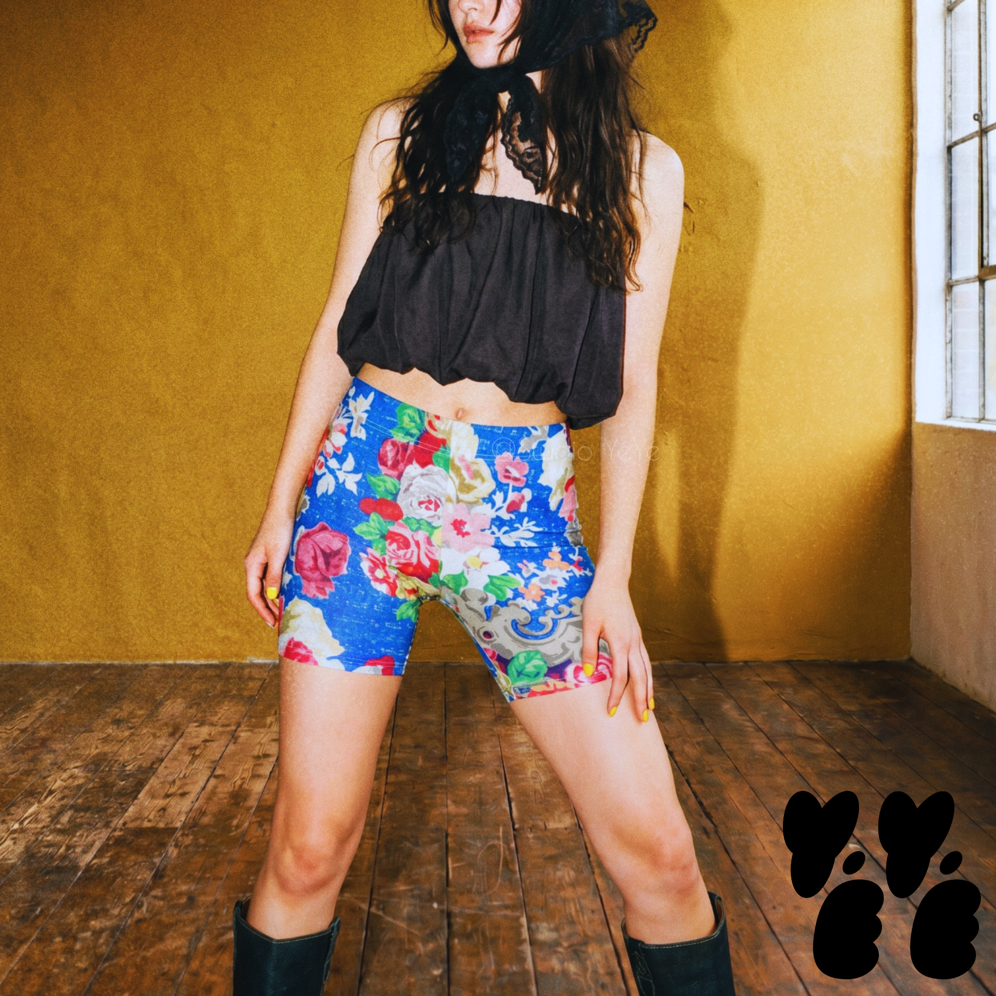 A female model wears 18th century rococo style floral printed bike shorts with vintage lace scarf on her face, black balloon shaped top and western boots 