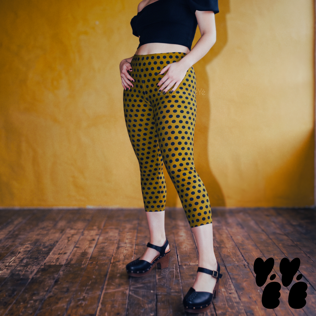 Model wearing dark mustard capri leggings with a navy blue polka dot pattern, styled with a black top and Mary Jane heels.