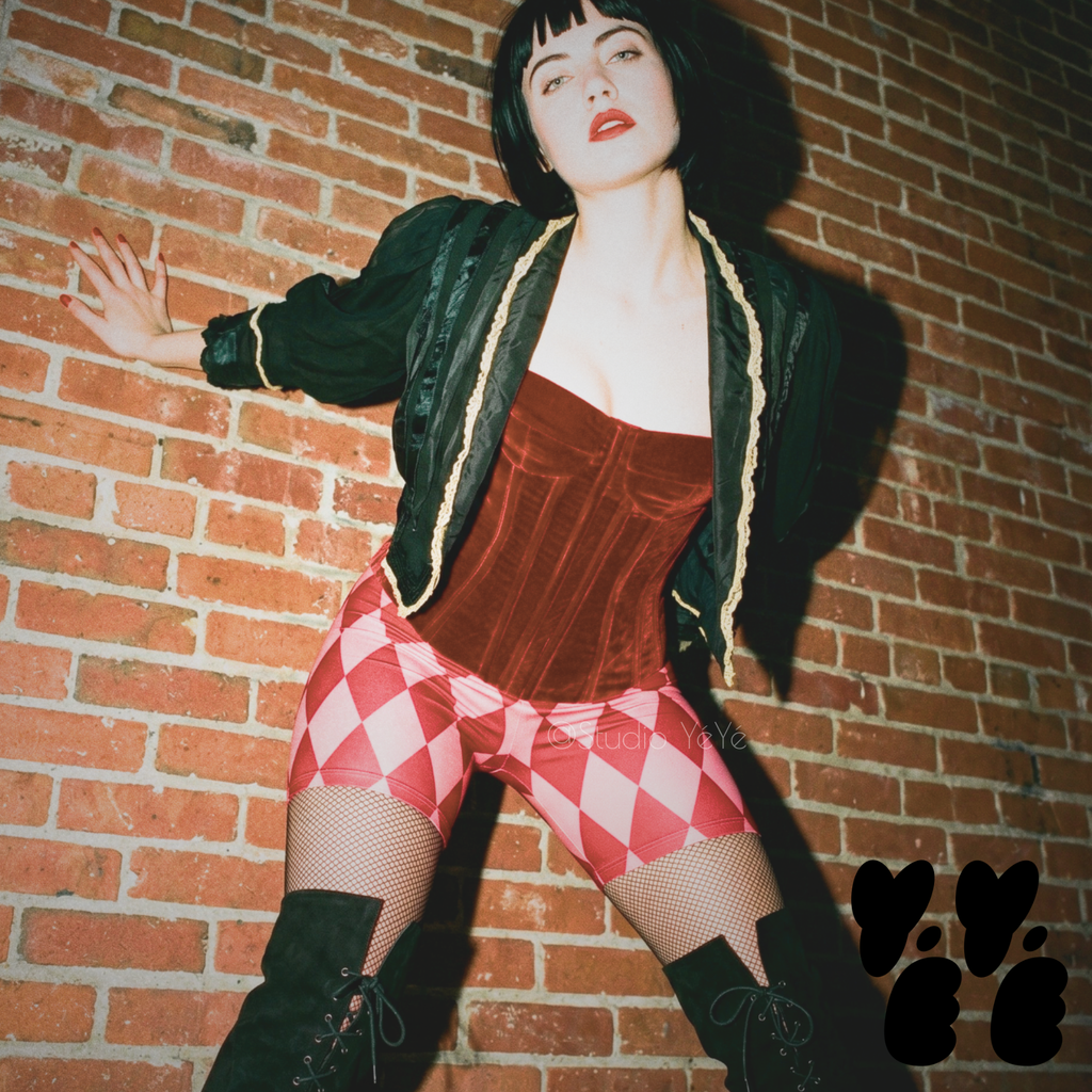 A female model wears harlequin diamond print bike shorts in red and pink, hipster edgy vibe, with vintage clothes.
