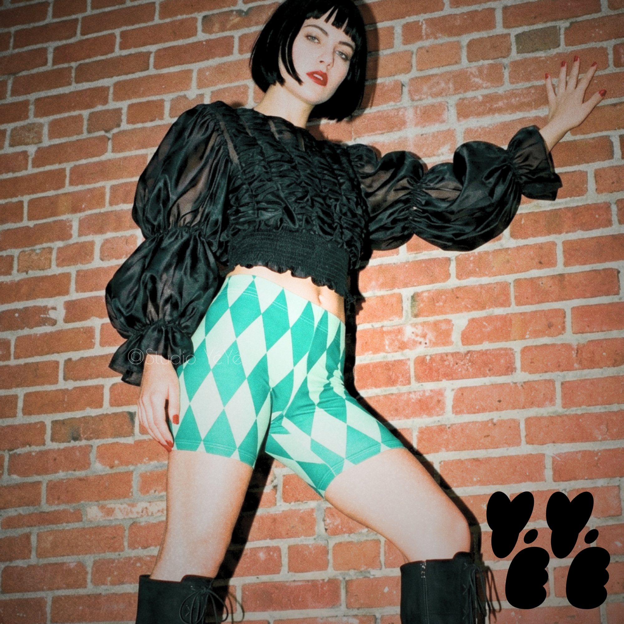 Teal and mint green vintage harlequin diamond bike shorts by YéYé, styled with a black ruffle puff sleeve crop top and black lace-up knee-high boots against a brick wall