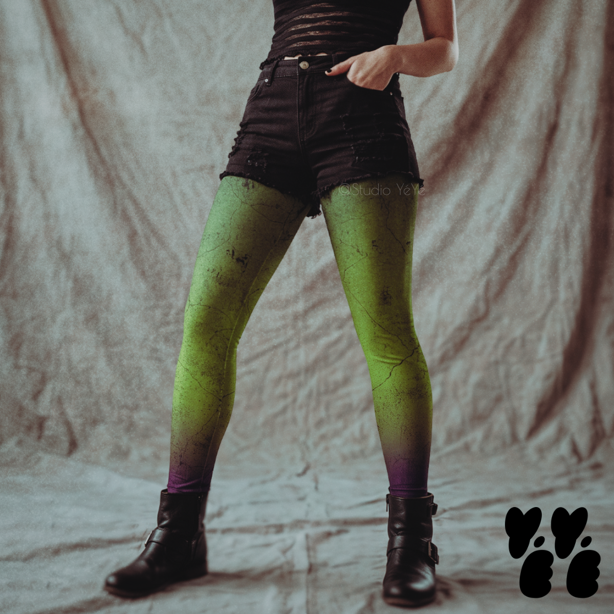 Model wearing toxic green leggings with a cracked, weathered wall texture fading to purple at the ankles, styled with black shorts and boots.