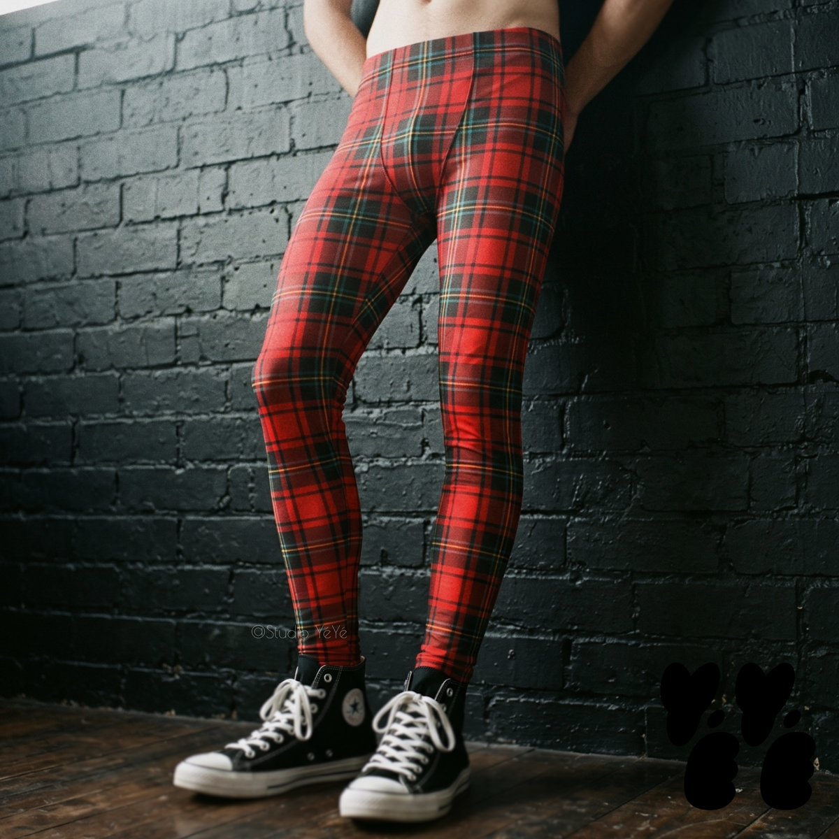 Model wearing red and black tartan plaid leggings with black high-top sneakers against a black brick wall.