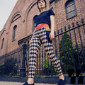 Model posing in front of a Brooklyn brick building in black and cream harlequin diamond leggings, featuring a red waistband and mustard yellow pocket details.