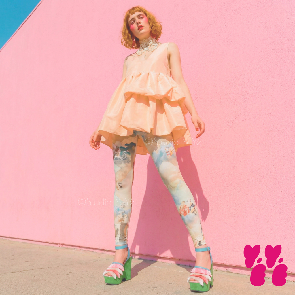 Renaissance angel cloud print leggings by YéYé in soft blue and cream, styled with a peach ruffle peplum top, pearl collar necklace and green platform sandals against a pink wall