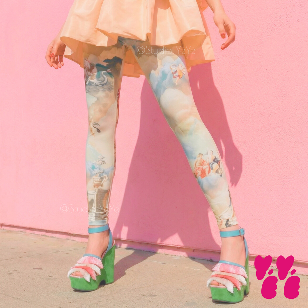Renaissance angel cloud print leggings by YéYé in soft blue and cream, styled with a peach ruffle peplum top, pearl collar necklace and green platform sandals against a pink wall